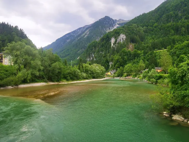 BookNoFees tatil evi: Hieflau, Styria, Avusturya – Ormanlık dağların ve kıyıda bulunan sarı bir evin yer aldığı nehir manzarası